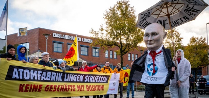 Foto: mehrere Menschen demonstrieren vor den Emslandhallen in Lingen und halten ein Banner, mit dem sie die Schließung der Brennelementefabrik fordern. Daneben eine übermannshohe Putin-Figur mit Rosatom-Shirt und unter einem ANF-Schirm