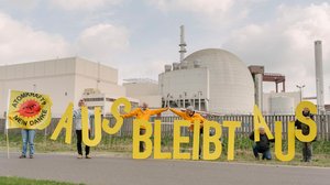 Aktivist*innen vor dem abgeschalteten AKW Brokdorf. Sie halten gelbe Schilder mit der Aufschrift „Aus bleibt aus!“ hoch. Eine Person schwenkt eine Fahne mit der Anti-Atom-Sonne.