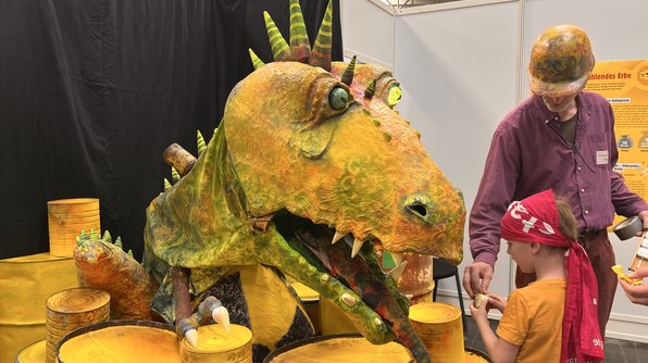 Foto: Ein Junge und ein Mann spielen mit einem übermannsgroßen Dinosaurier aus Pappmaché